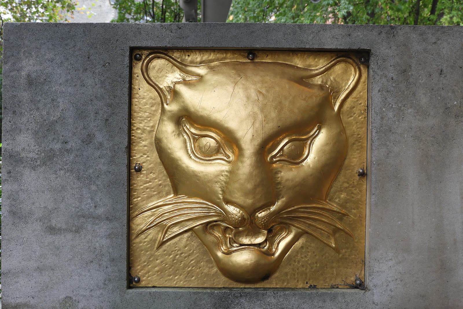 A gilded relief of a leopard's head with whiskers and bared teeth, set into a grey stone pillar